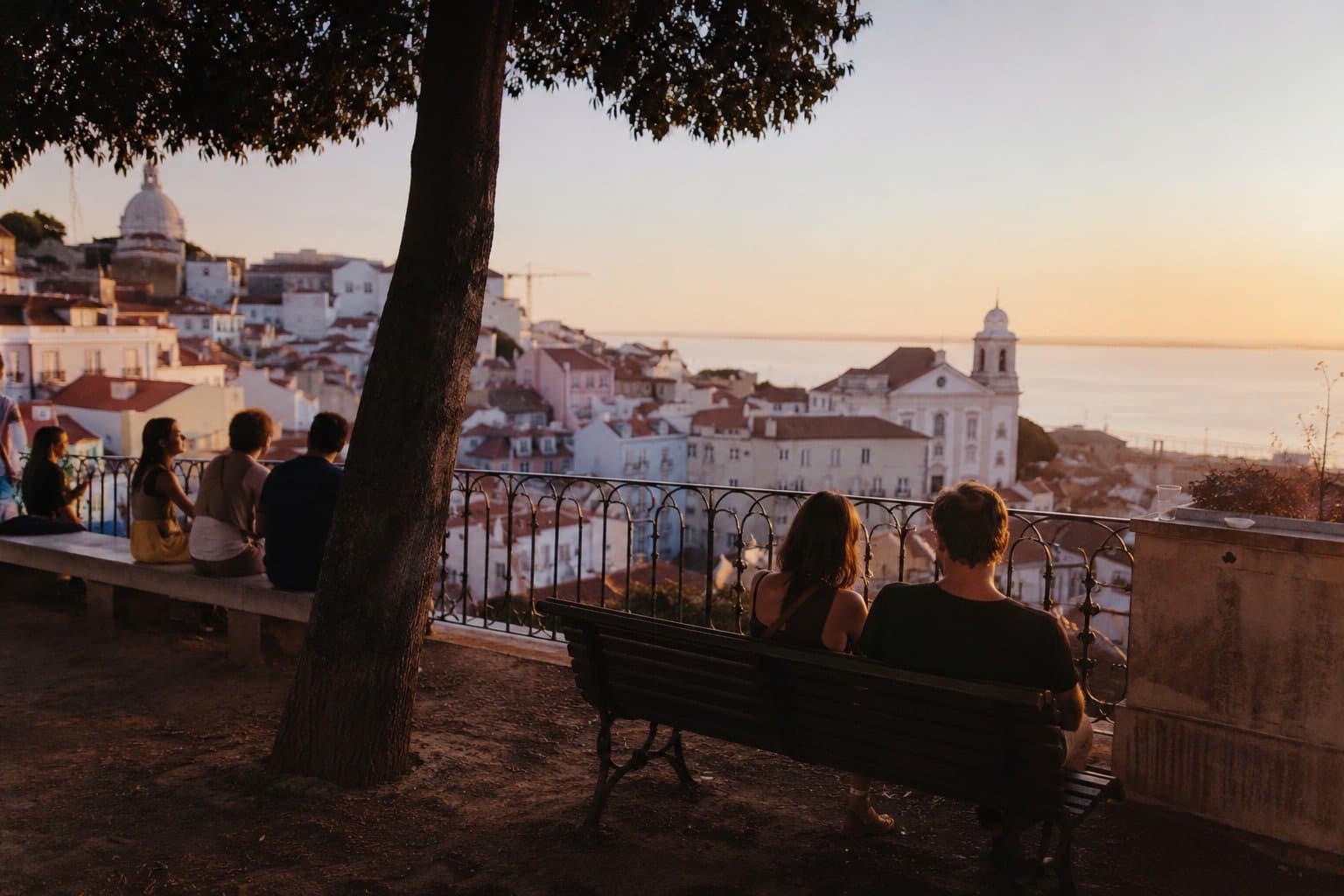 Gente sentada viendo el atardecer sobre los tejados de Alfama desde un miradouro, Lisboa